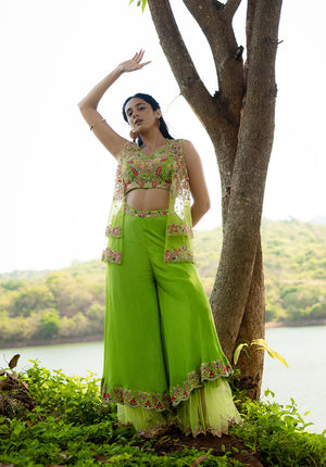 Model wearing a green sharara set with matching blouse, sharara pants, and layered embroidered jacket, posing near a tree.