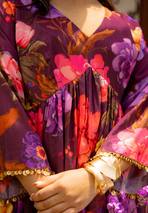 Close-up of V-neck purple floral dress featuring golden sequin trim sleeves and gold bangles