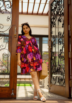 Model standing in doorway wearing purple floral short dress with white heels and straw bag
