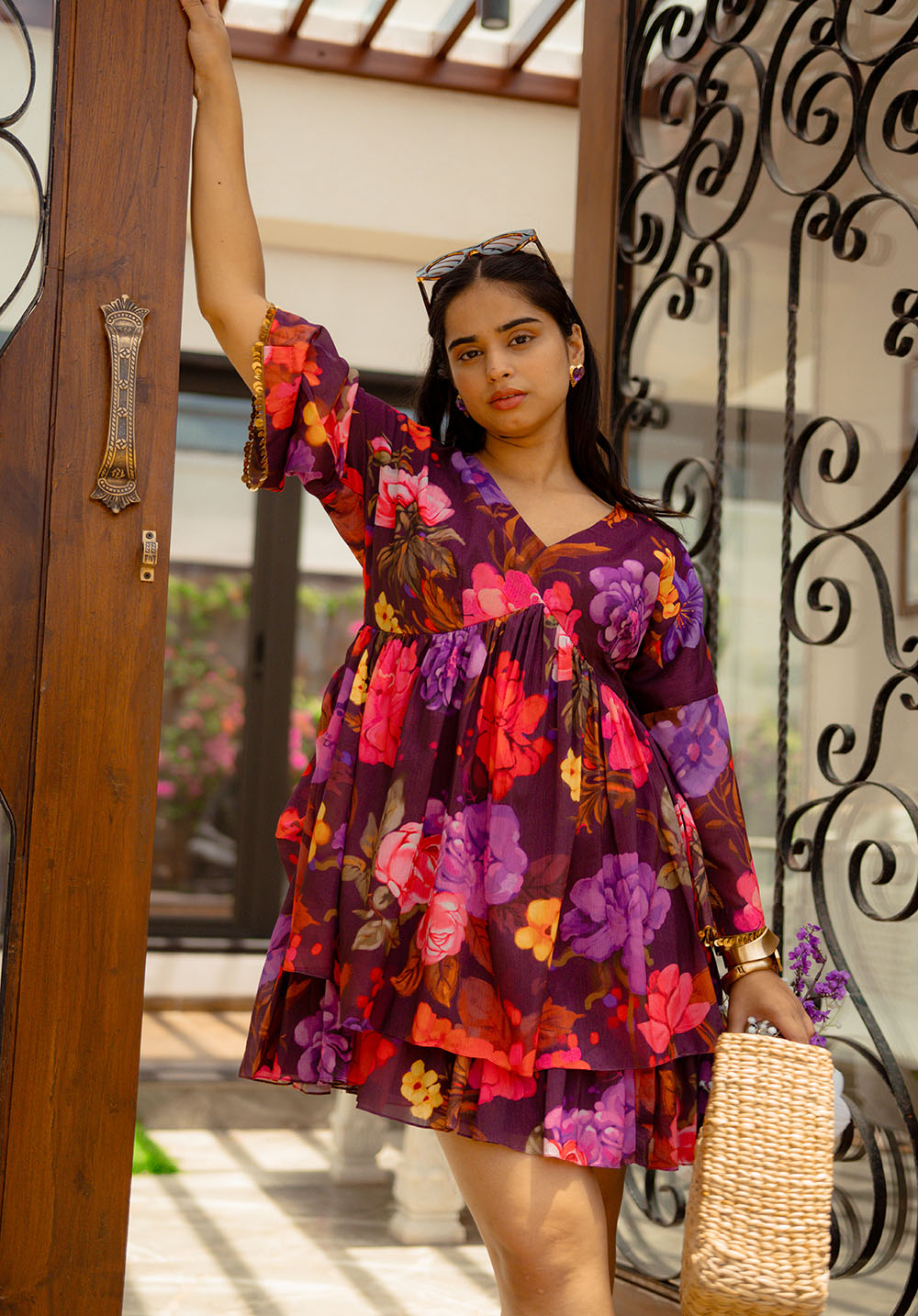 Model standing and posing in a purple floral dress with gold sequin trim, holding a straw handbag
