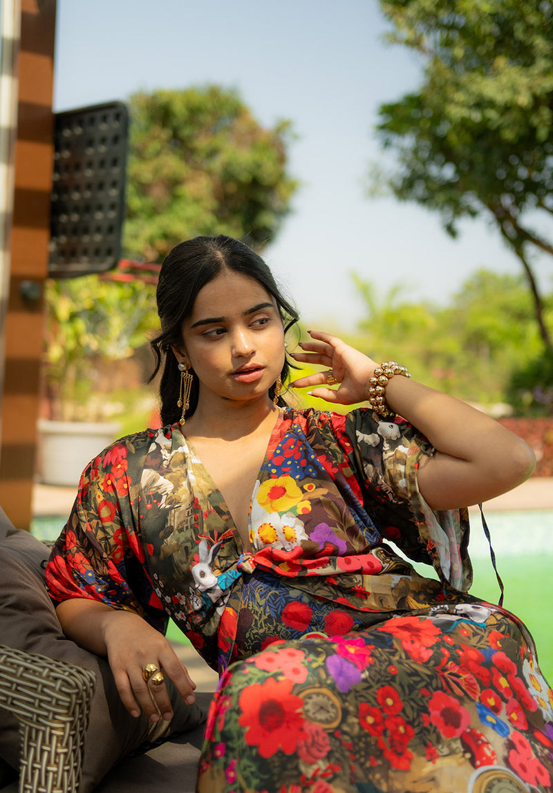 Model seated on a chair wearing a floral dress with V neckline and relaxed sleeves, accessorized with a golden ring
